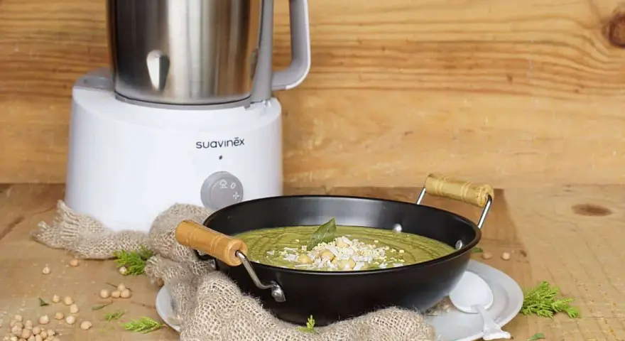 Un puré verde cremoso, ideal para nabo bebés, servido en un cuenco negro con garbanzos y hierbas. Al fondo, una máquina de cocina Suavinex.