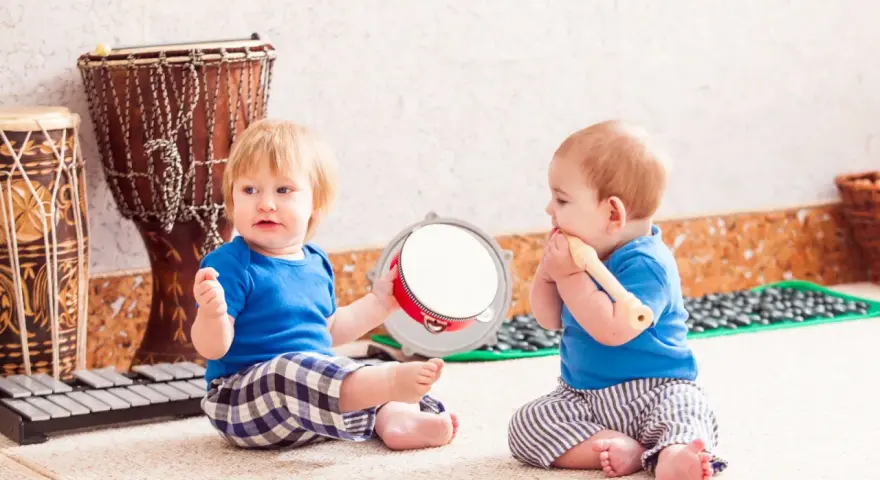 Dos bebés disfrutan de la estimulación musical temprana, uno con un tambor y otro con una flauta, rodeados de instrumentos.