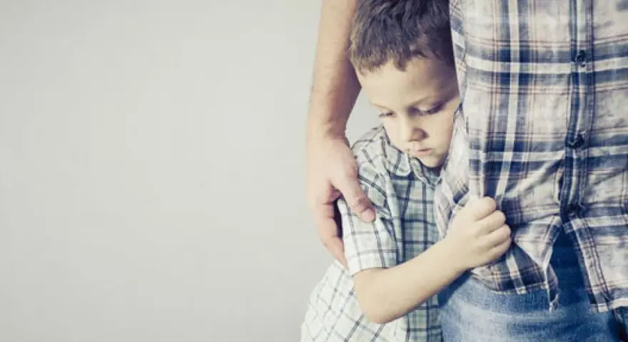 Un niño se aferra a la pierna de un adulto, con la cabeza baja, reflejando una **crisis** de **8 meses**.