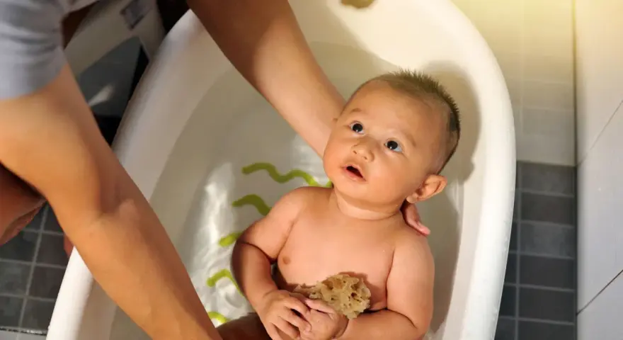 Bebé en bañera, manos adultas ajustan la temperatura del agua. Sostiene una esponja, mirando hacia arriba con curiosidad.