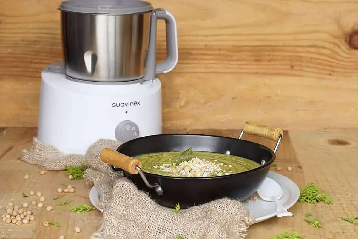 Un puré verde cremoso, ideal para nabo bebés, servido en un cuenco negro con garbanzos y hojas de laurel. Al lado, una máquina de cocina Suavinex.