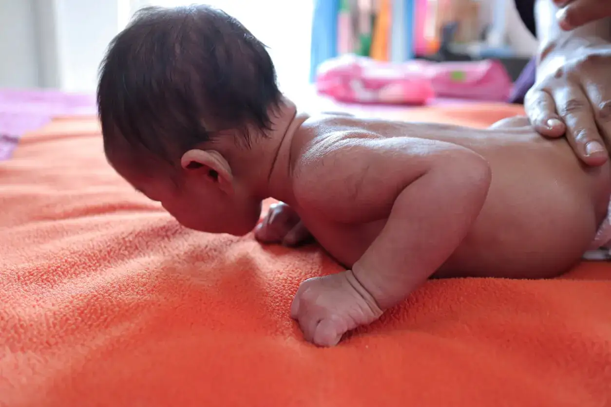 Un bebé boca abajo sobre una manta naranja, con una mano adulta sujetando su espalda. Es importante saber porque hay que sujetar la cabeza a los bebés.