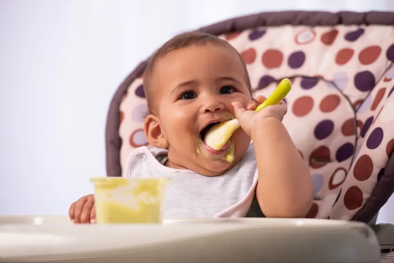 Beb&eacute; feliz probando su primer pure para bebes con una cuchara verde.