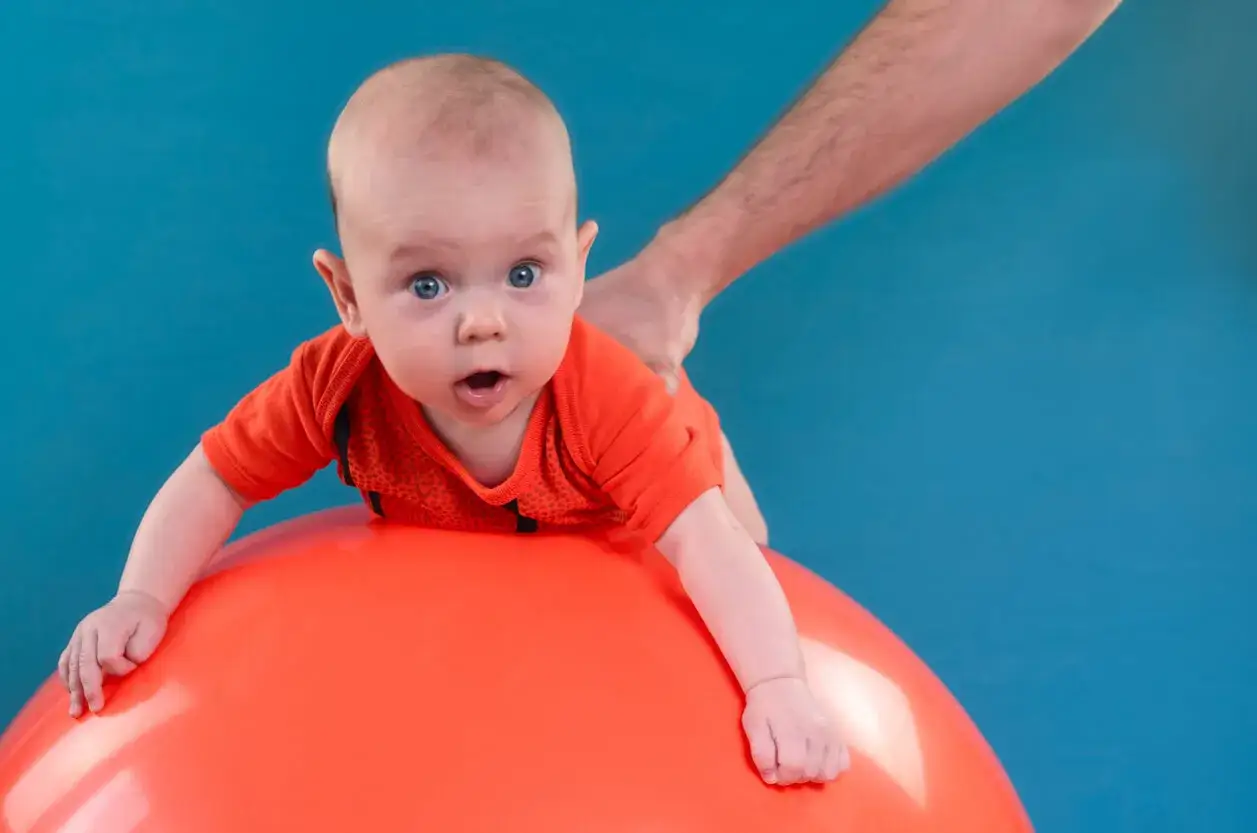 Bebé boca abajo sobre una pelota naranja, con una mano adulta sujetando su espalda. Es importante sujetar la cabeza a los bebés para su desarrollo.