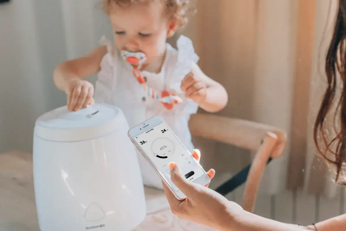 Un beb&eacute; juega cerca de un humidificador, mientras una mano controla el aparato desde un m&oacute;vil. Este humidificador beb&eacute; para que sirve es ideal para crear un ambiente confortable.
