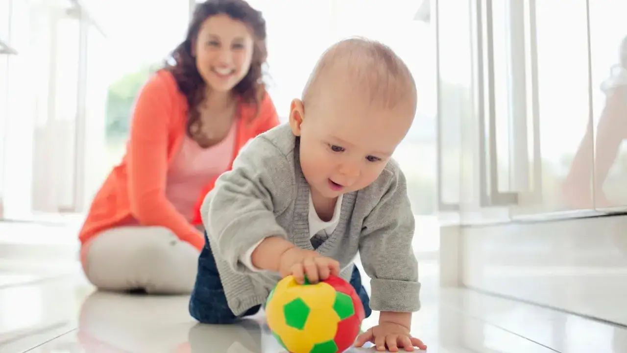 Beb&eacute; gateando hacia una pelota de colores, disfrutando del movimiento libre mientras su madre sonr&iacute;e al fondo.