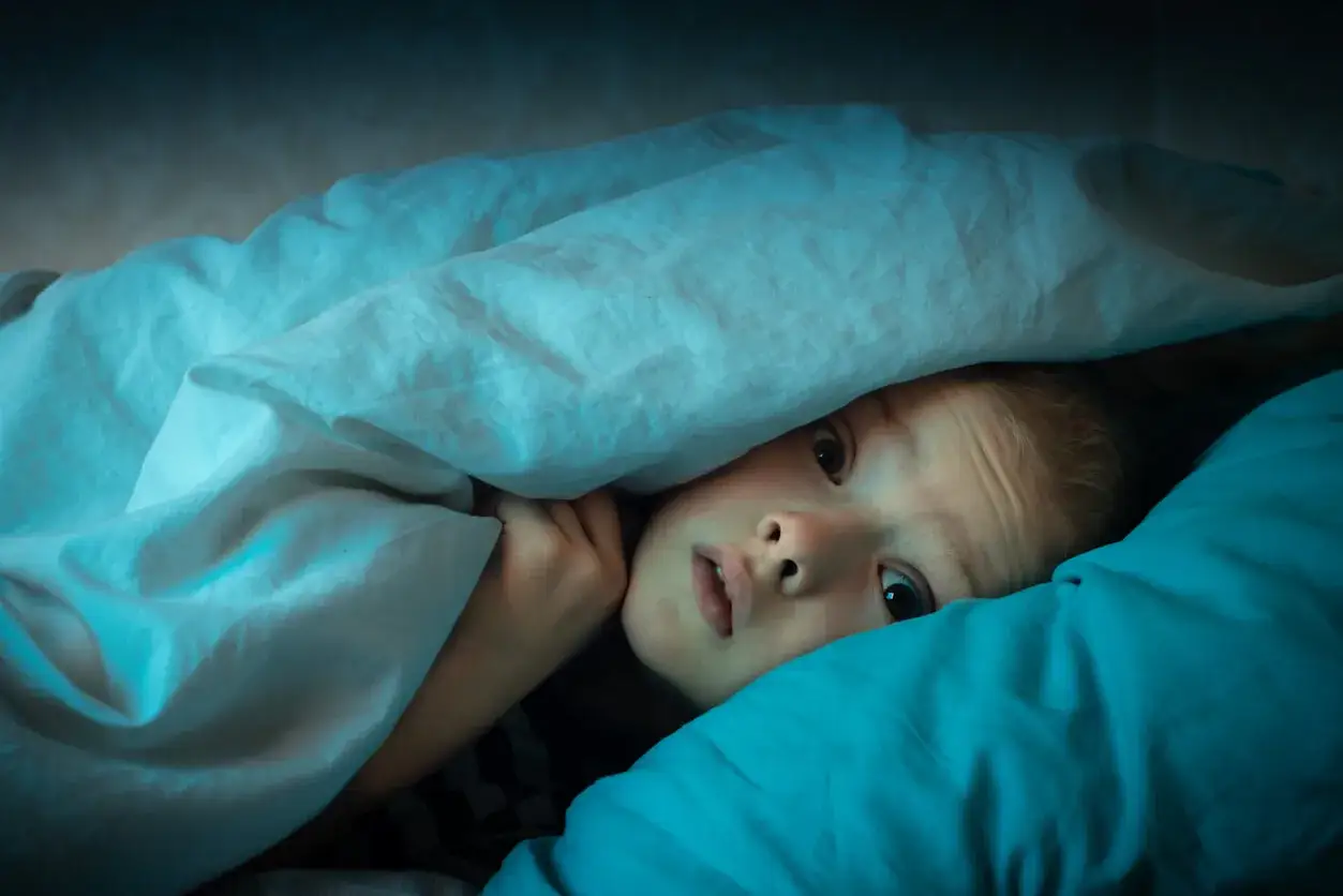 Un niño asustado se esconde bajo las sábanas, sus ojos reflejan los terrores nocturnos infantiles.