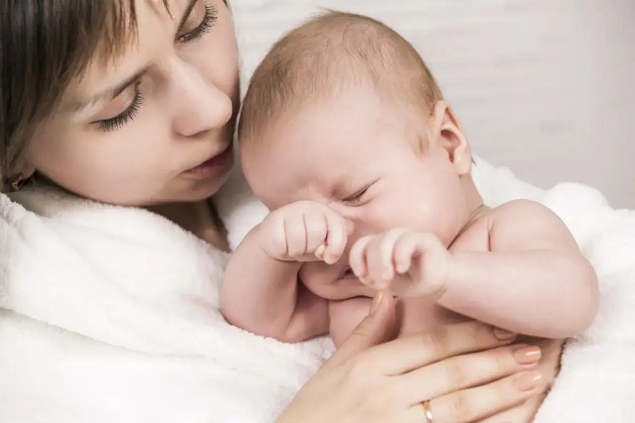 Madre reconforta a su bebé, que parece tener una crisis de 8 meses, acurrucados en una manta blanca.