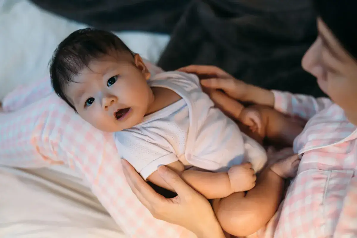 Beb&eacute; asi&aacute;tico despierto en brazos de madre, con boca abierta, listo para comer. La madre lo sostiene suavemente, un momento tierno de c&oacute;mo despertar a un beb&eacute; para que coma.