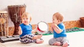 Dos bebés disfrutan de la estimulación musical temprana, uno con un tambor y otro con una flauta, rodeados de instrumentos.