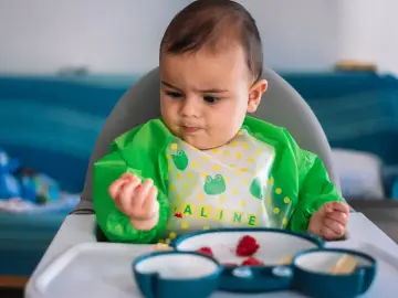 Bebé con babero verde, aprendiendo a comer con las manos. En su plato hay trozos de fruta.