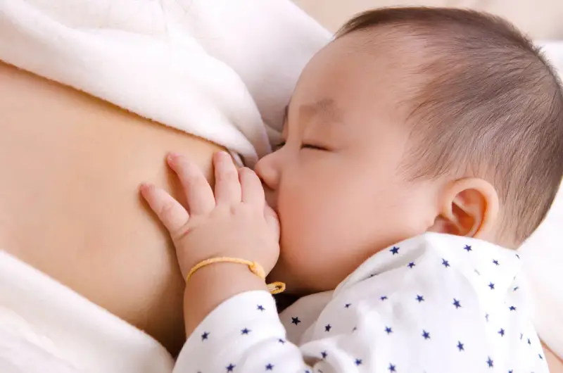 Bebé asiático durmiendo mientras se alimenta, mostrando el reflejo de succión. Su mano pequeña descansa sobre el pecho de su madre.