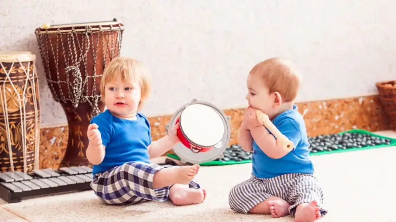 Dos bebés disfrutan de la estimulación musical temprana, uno con un tambor y otro con una flauta, rodeados de instrumentos.